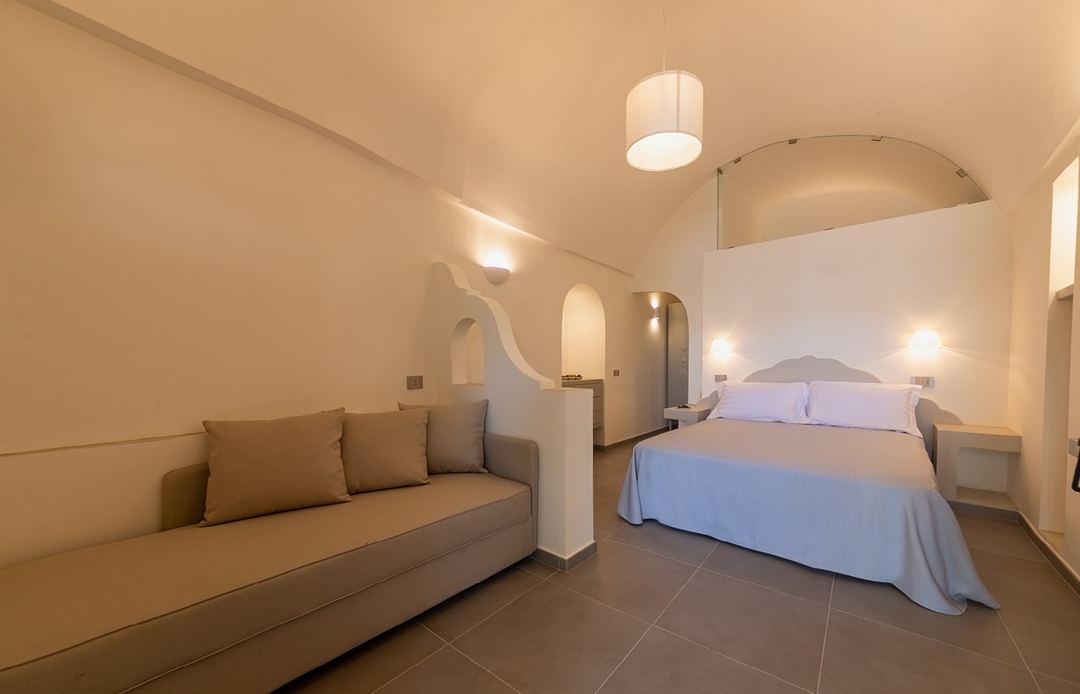 A modern boutique hotel room featuring a bed with blue linens, a tan sofa, arched architectural details, and warm ambient lighting.