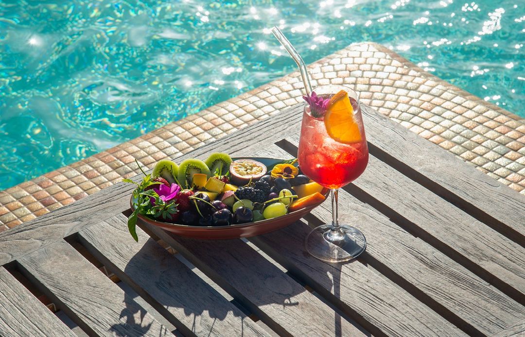 A refreshing tropical cocktail and fresh fruit platter sit poolside on a wooden deck overlooking crystal-clear turquoise water.