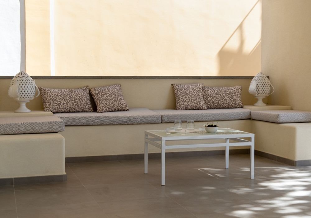 A minimalist lounge area featuring a neutral-toned built-in seating arrangement with patterned cushions, a white metal coffee table, and abundant natural light from a large window.