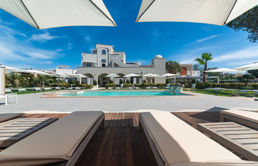 The hotel's turquoise swimming pool with white umbrellas and sun loungers overlooking the Mediterranean-style main building under a clear blue sky.