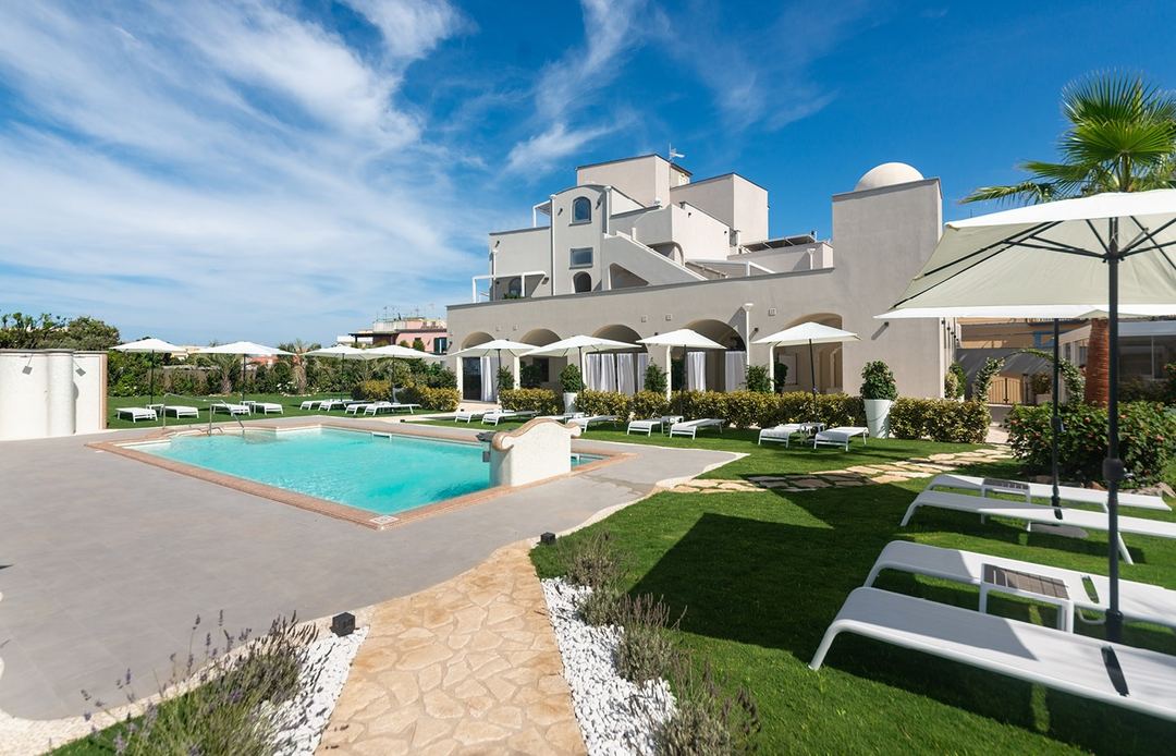 The hotel's pool area features a bright turquoise swimming pool surrounded by white umbrellas, lounge chairs, manicured lawns, and the whitewashed main building under a clear blue sky.