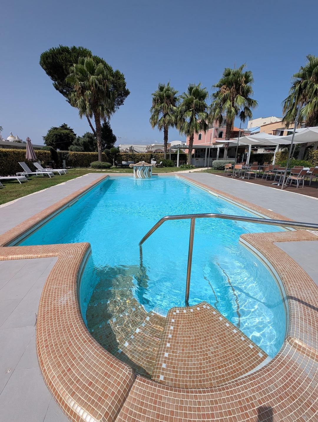 A curved spa pool in the foreground with an Olympic-length swimming pool beyond, surrounded by manicured lawns, palm trees, and a Mediterranean-style building under clear blue skies.