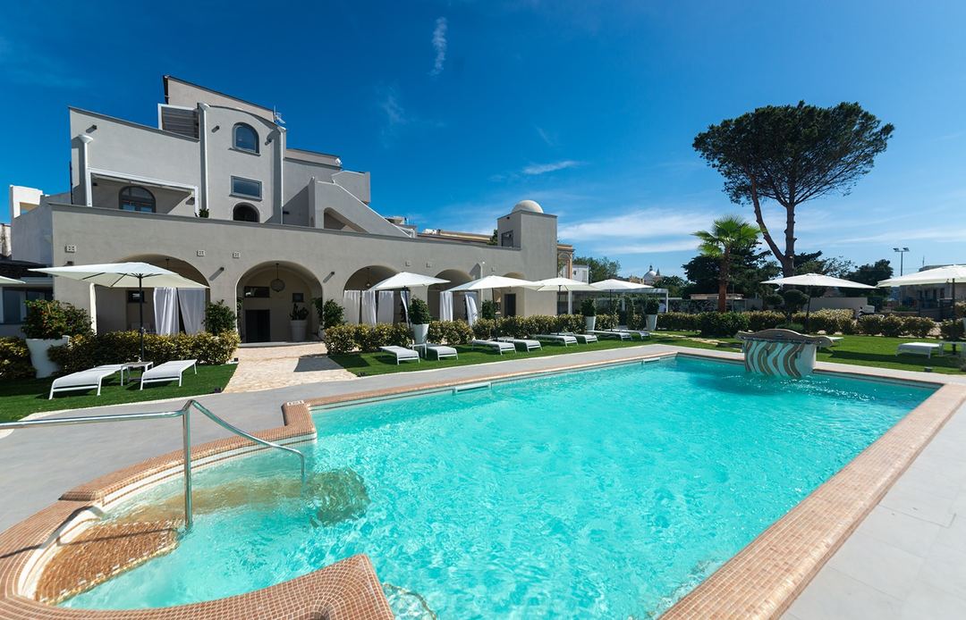 The Insula Boutique Hotel's swimming pool featuring crystal-clear turquoise water, surrounding loungers and umbrellas, manicured lawns, and the whitewashed architecture of the hotel building under a clear blue sky.