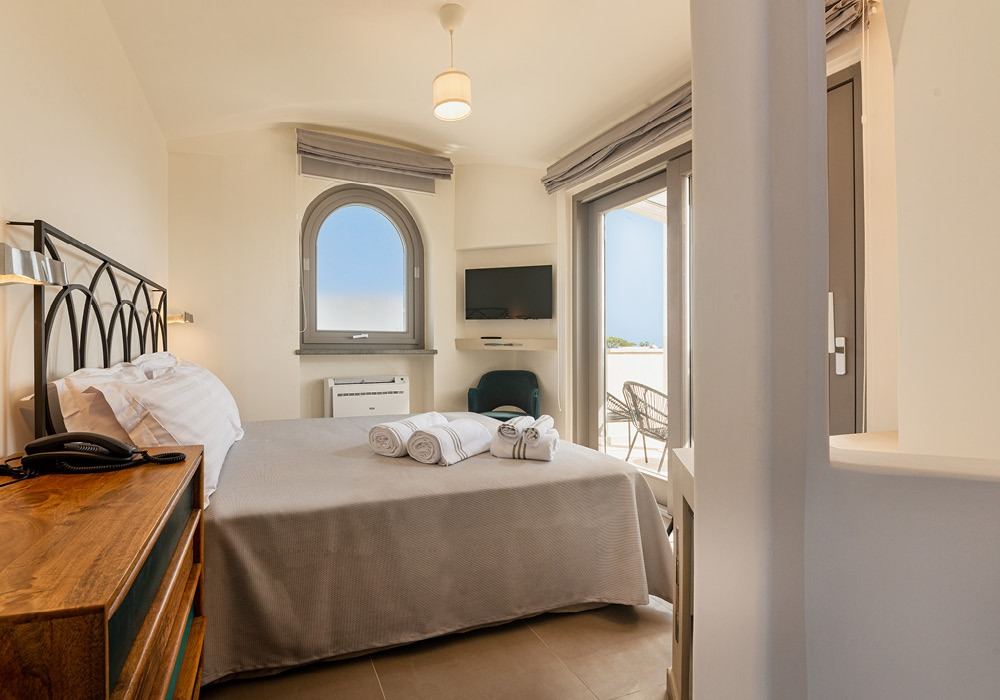 A bright bedroom with a double bed, arched window with sea views, flat-screen TV, and access to a private balcony.