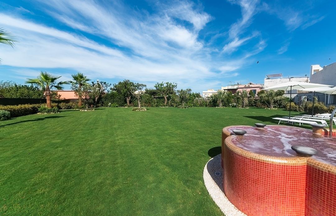 The hotel's expansive manicured gardens feature a red-tiled hot tub, palm trees, and sun loungers under a clear Mediterranean sky.